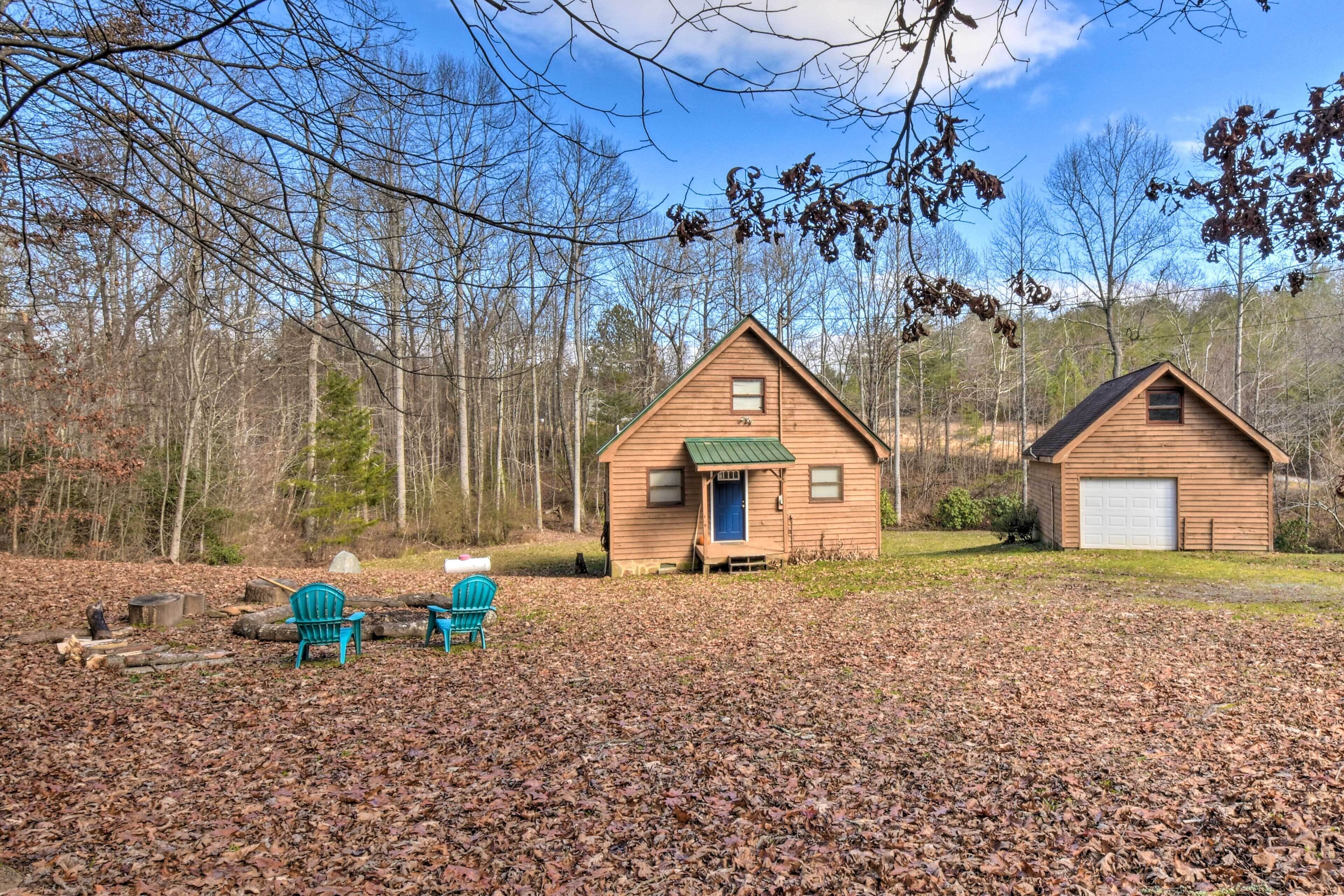A cabin with a green roof and a garage, surrounded by trees and a leaf-covered ground.