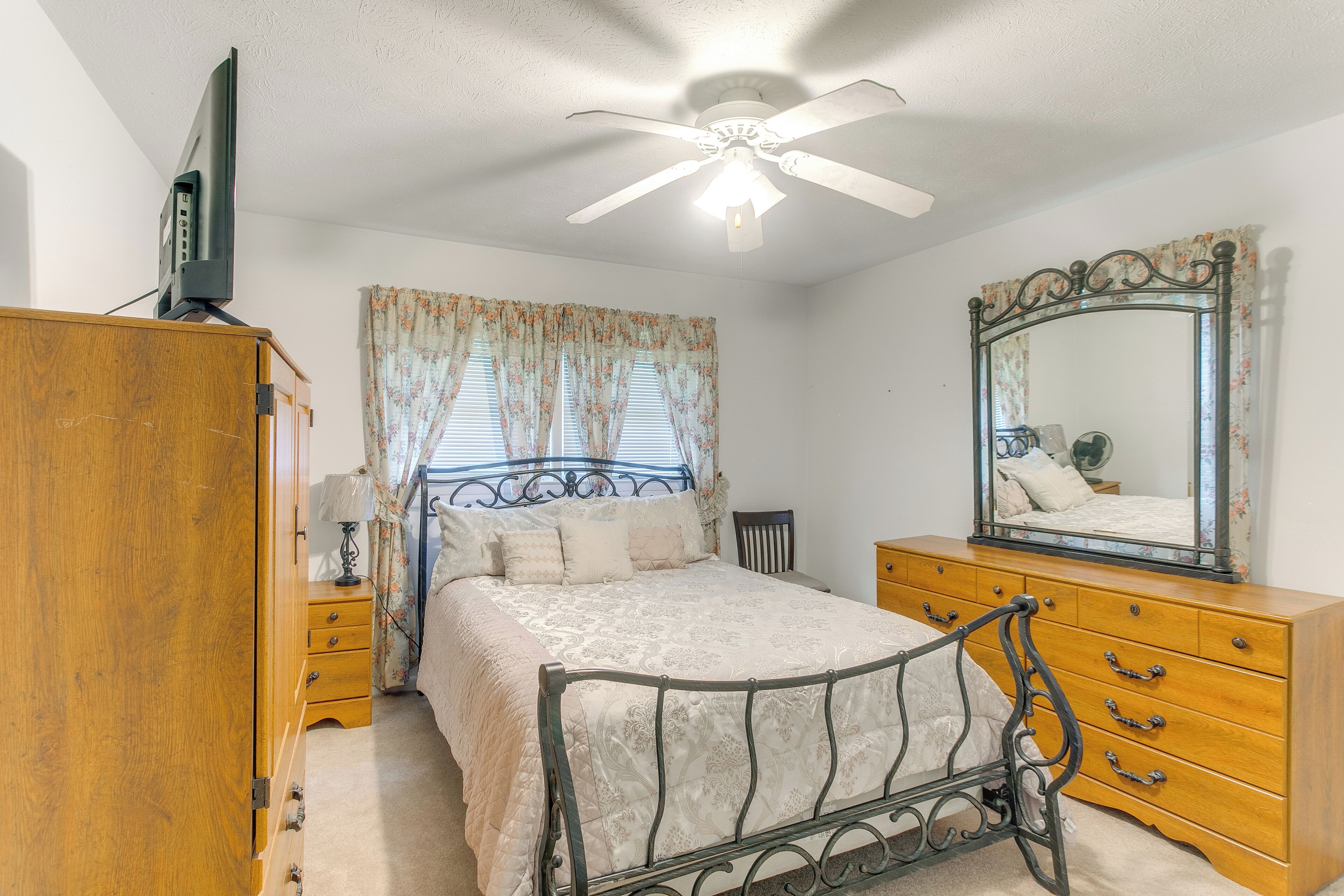 A bedroom with a bed, wooden dresser, mirror, ceiling fan, and a television on a cabinet.