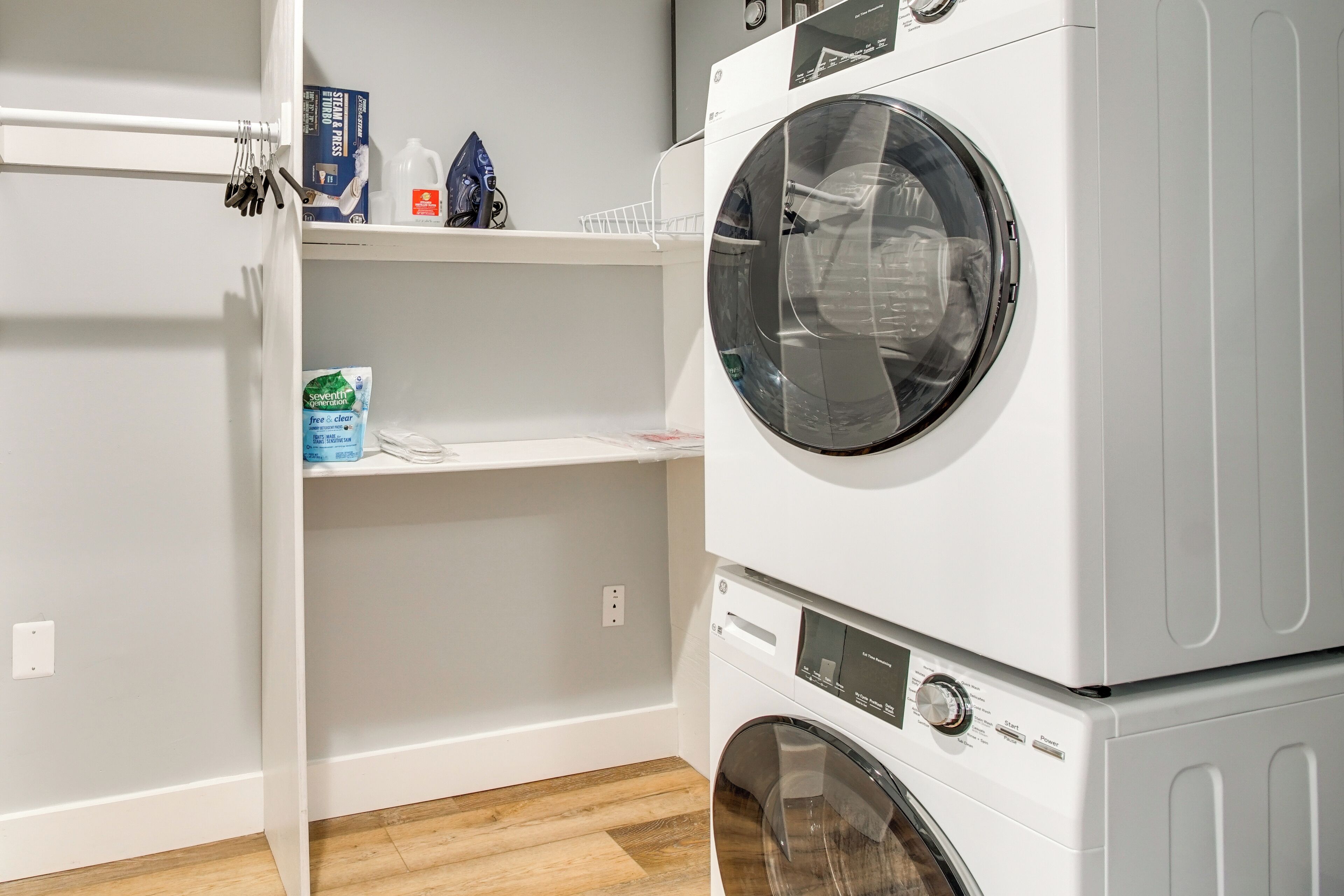 A laundry area with stacked washers and dryers, a shelf with detergent, and a rack with hanging clothes.
