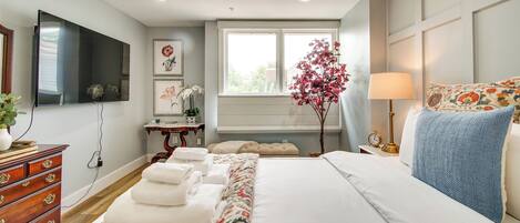 A hotel room with a large bed, a flat-screen TV mounted on the wall, a window with a view, and a bedside table with a lamp.