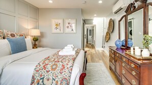 A well-lit bedroom with a large bed, a dresser, a mirror, and framed artwork on the wall.