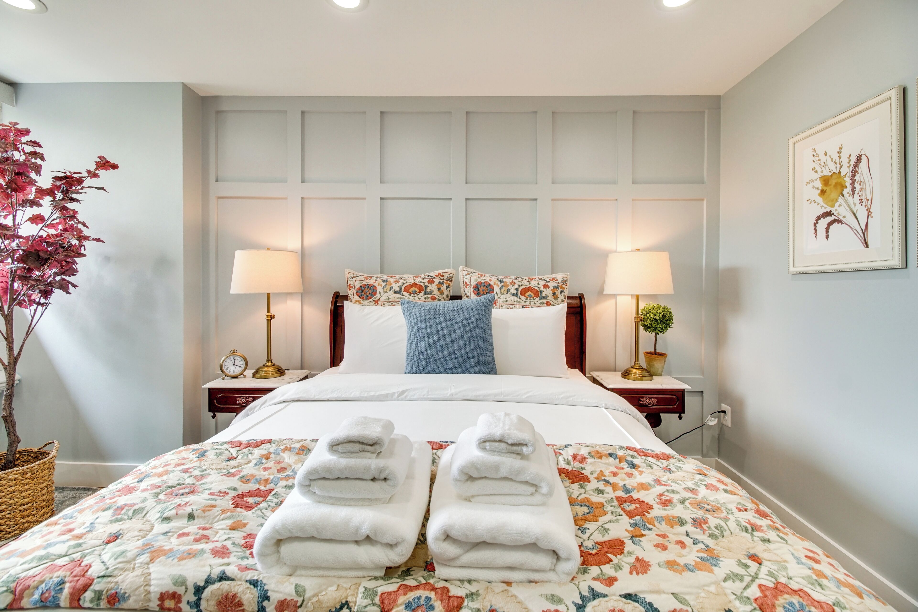A neatly made bed with a floral bedspread, two bedside lamps, a clock, and a framed floral artwork on the wall.