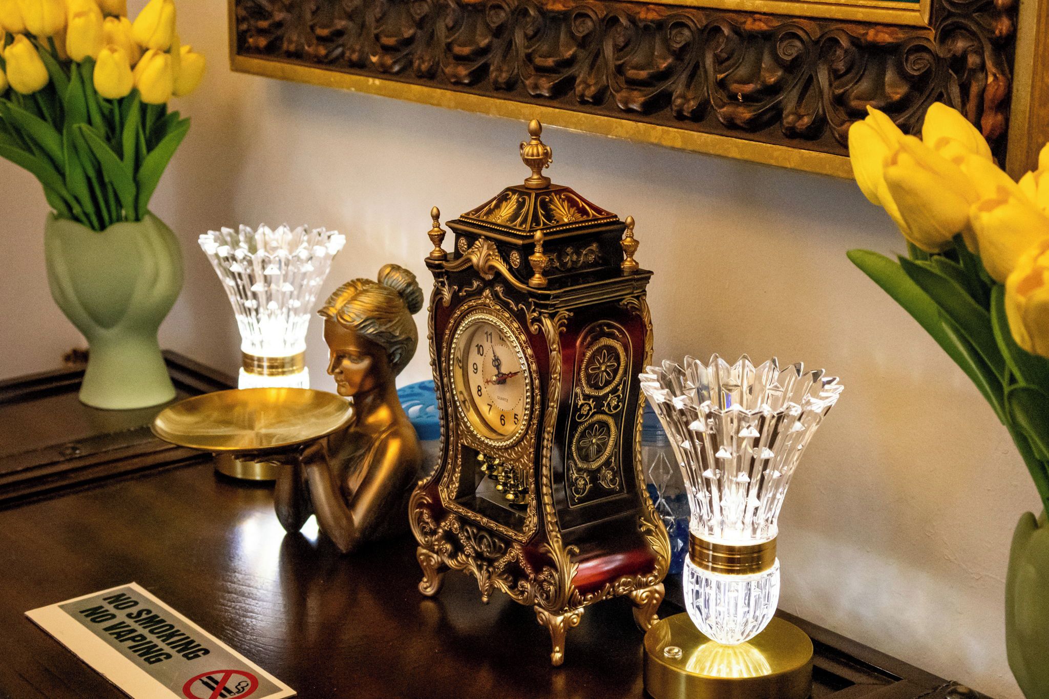 Une table ornée d’une horloge décorative, de deux vases en cristal et d’un bouquet de tulipes jaunes.