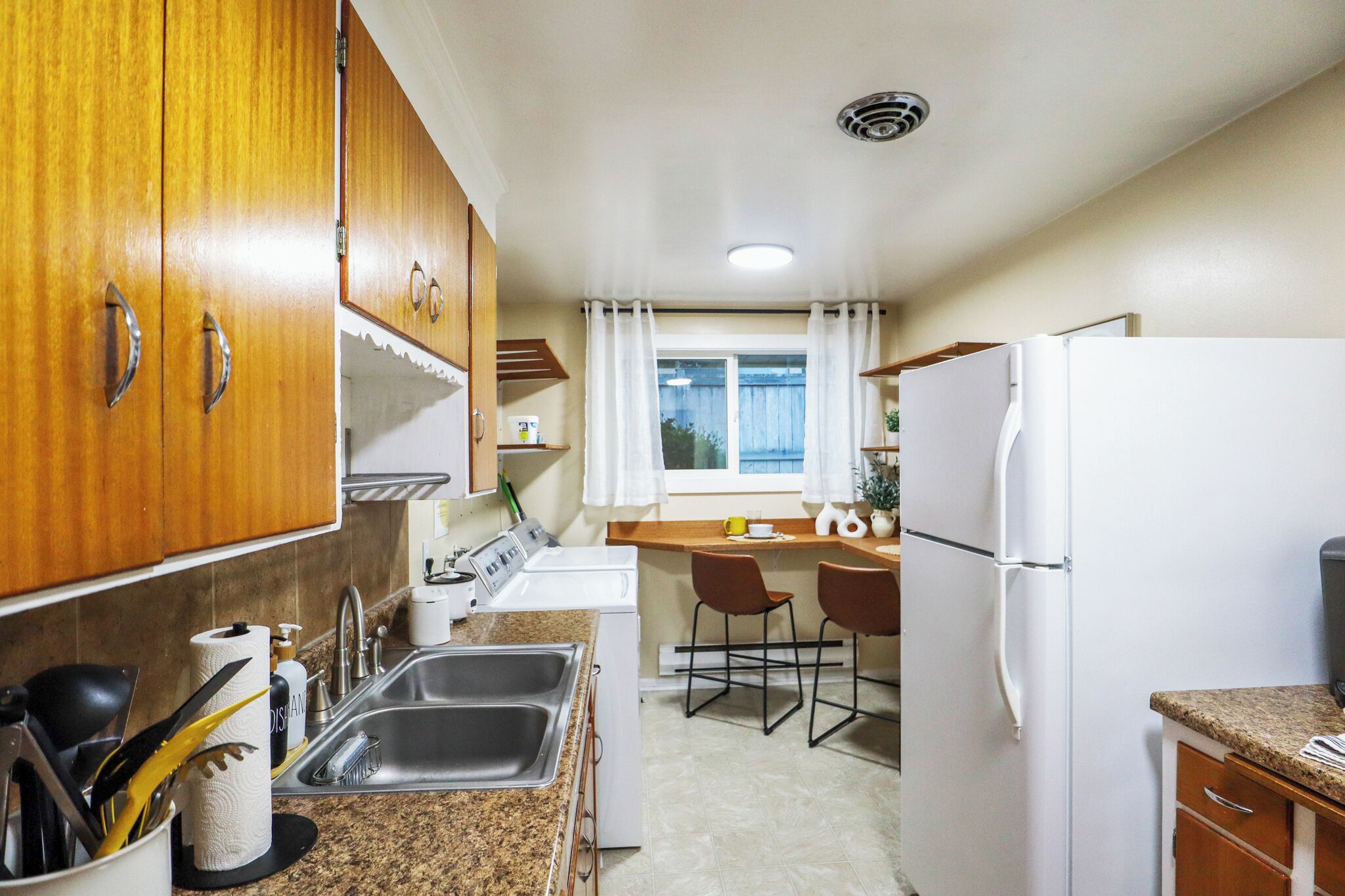 Cocina compacta con armarios de madera, un lavamanos, un refrigerador blanco y una ventana con cortinas.
