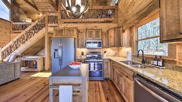 Interior rústico de una cabaña con cocina, escalera de madera y un amplio ventanal con vistas a un bosque.