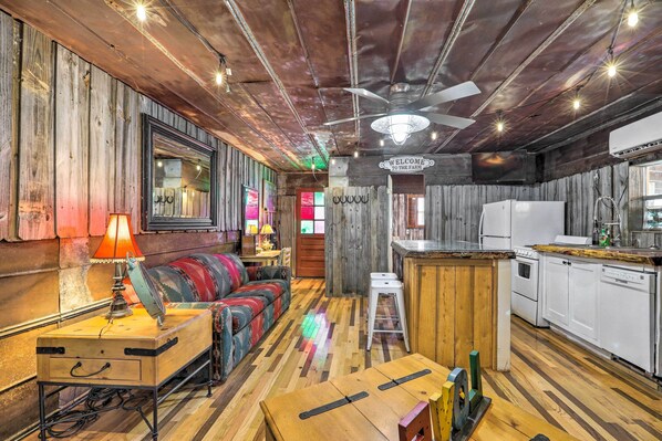 Un intérieur rustique avec un plafond en bois, un canapé, une table avec une lampe, un coin cuisine et une pancarte indiquant « Bienvenue à la ferme ».