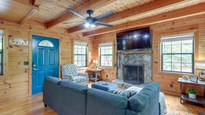 A cozy cabin interior with a blue door, a fireplace, a ceiling fan, and a large window.