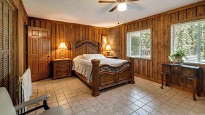 A bedroom with a wooden paneled wall, a bed with a headboard, two bedside lamps, a ceiling fan, and a window with blinds.