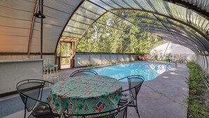 A covered outdoor area with a pool, a table with chairs, and a view of trees.