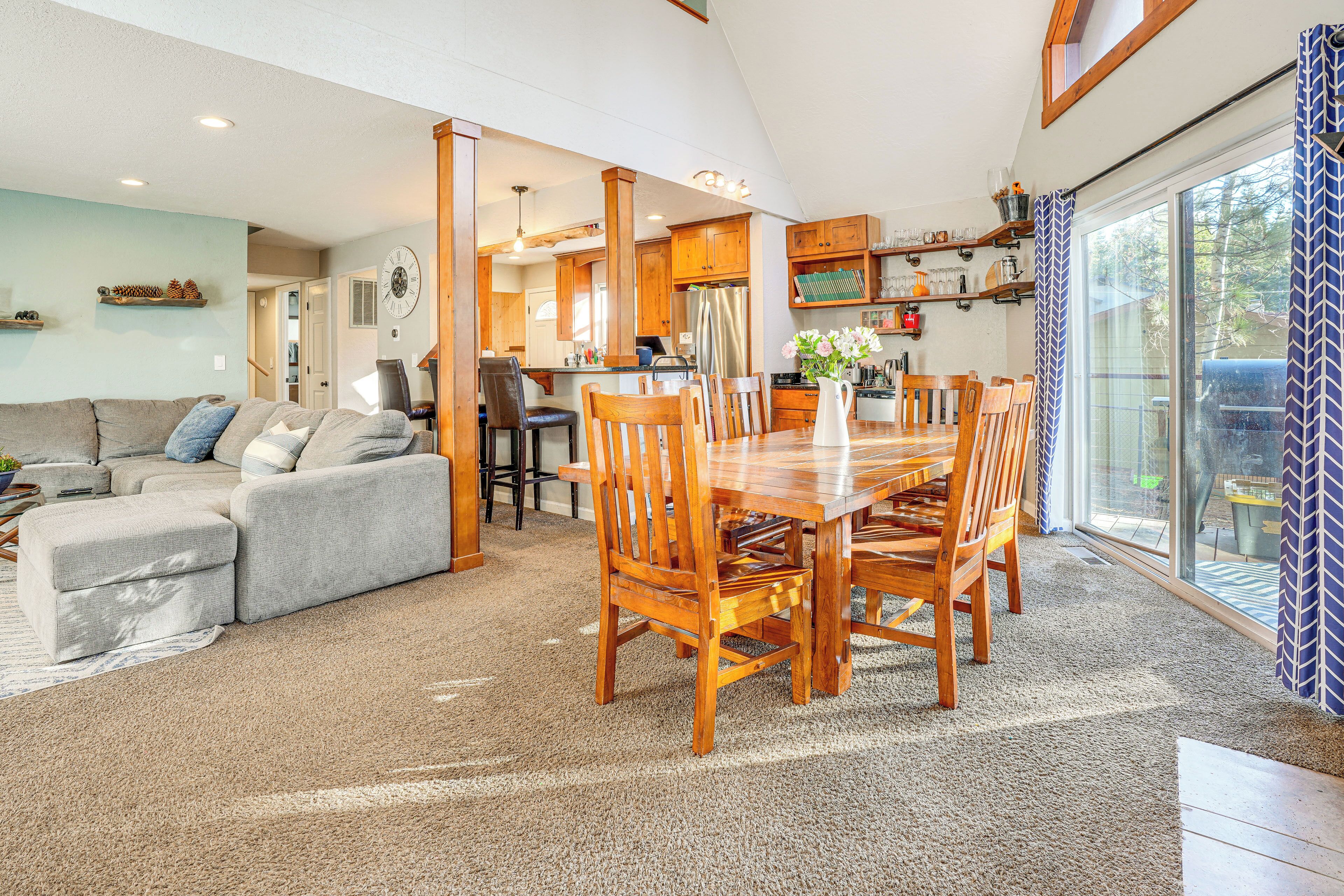 A spacious living area with a large wooden dining table, a grey sectional sofa, and a kitchen in the background.