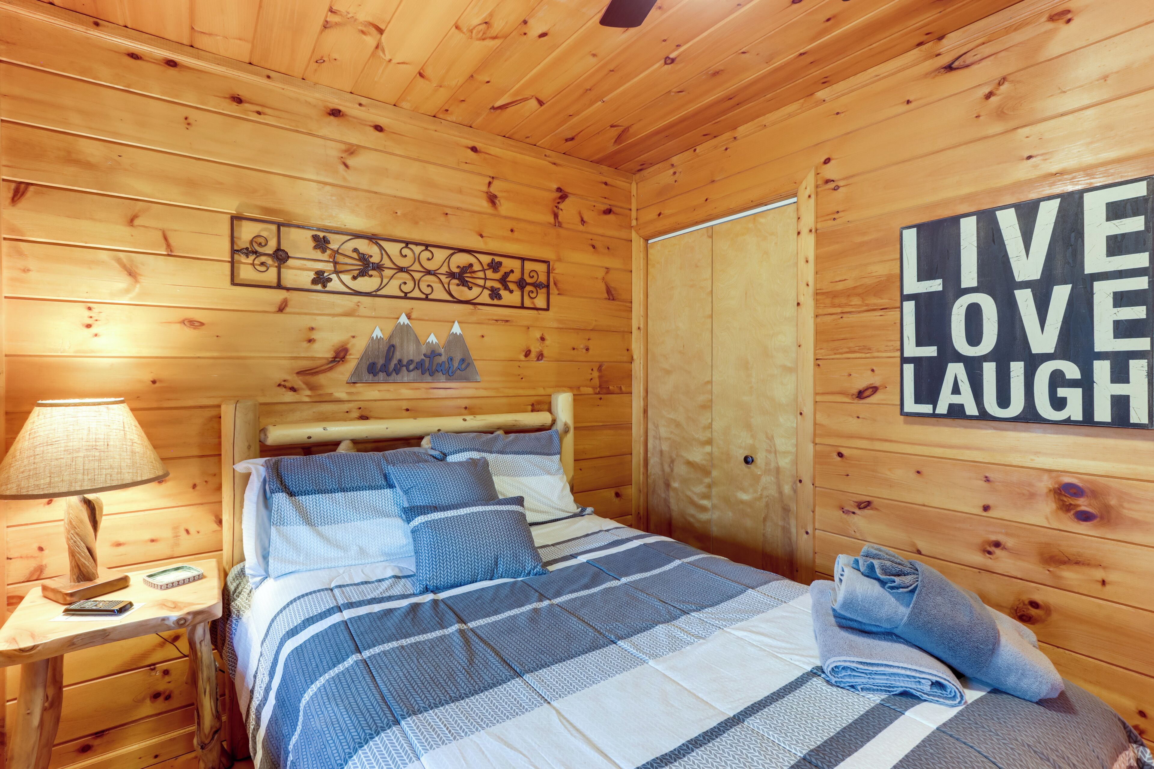 Un dormitori de cabana de fusta amb un llit, una tauleta de nit, una làmpada i un cartell al mur amb la frase "LIVE LOVE LAUGH".