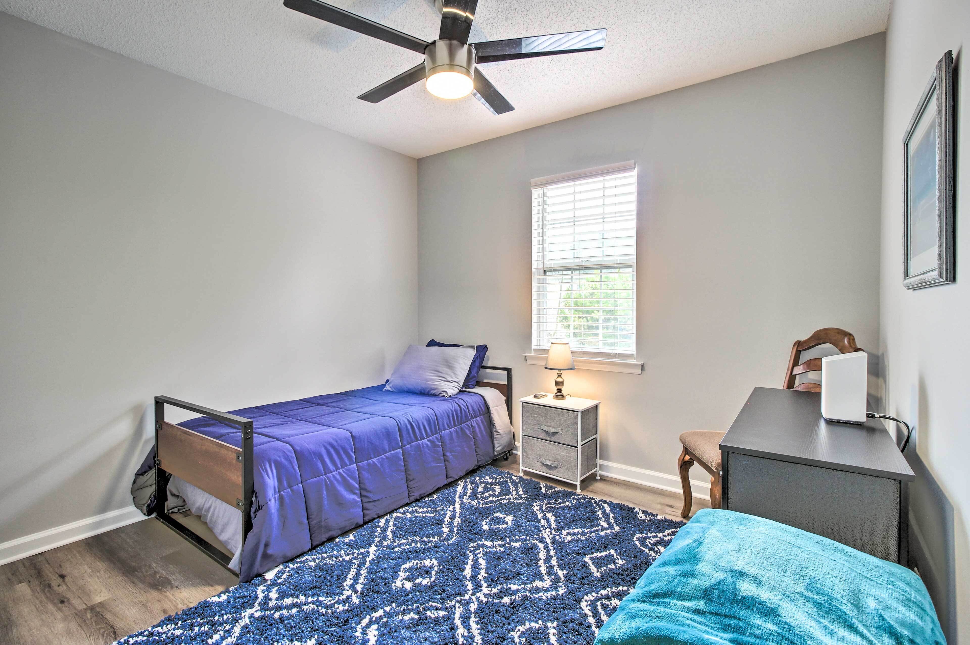 A bedroom with a bed, a desk, a chair, a nightstand, a lamp, a ceiling fan, and a window.