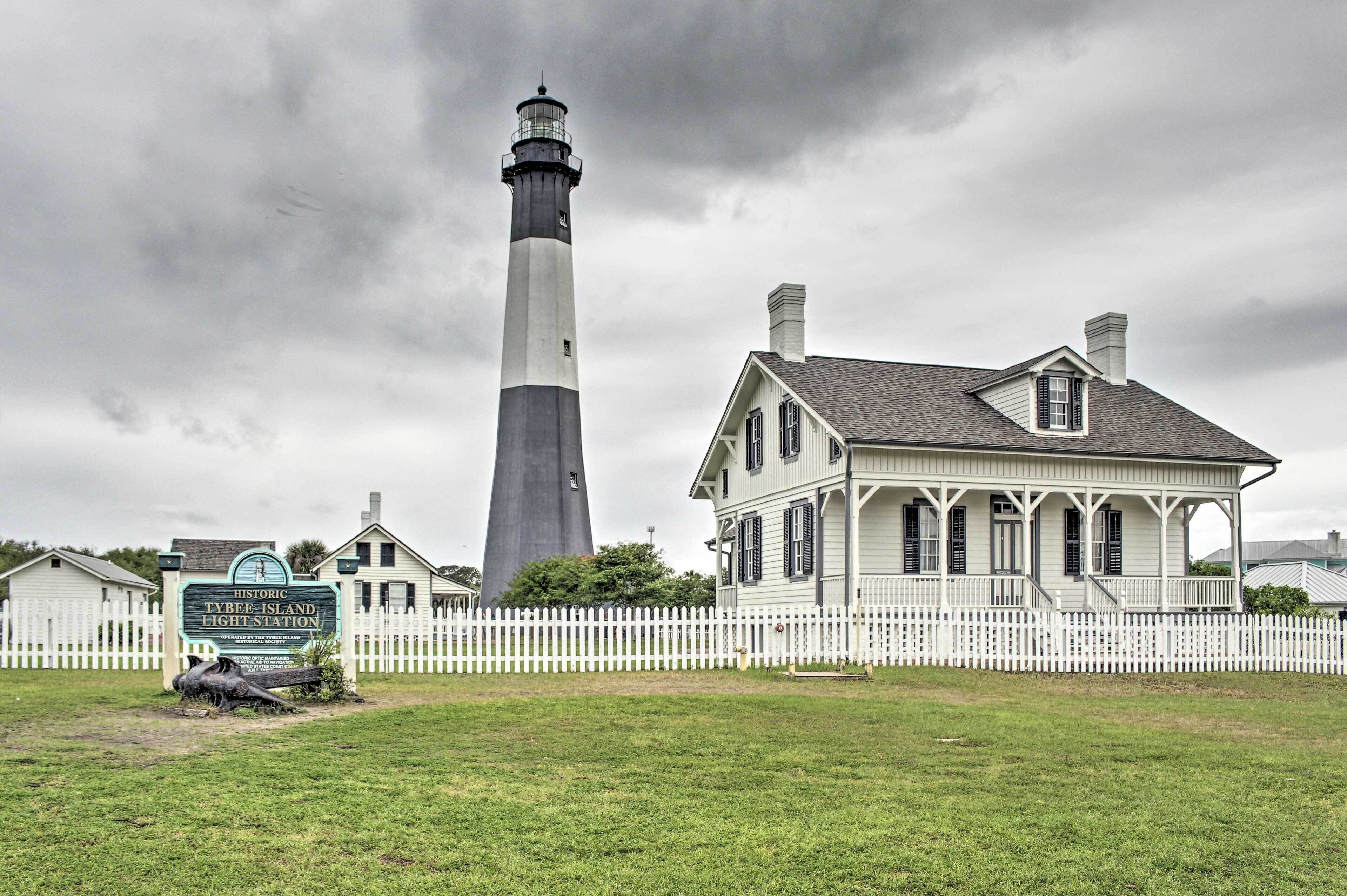 A historic lighthouse with a white and black tower, a white picket fence, and a historic building with a porch.