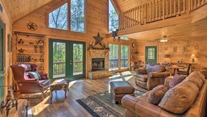 A spacious cabin interior with a stone fireplace, leather furniture, and large windows offering views of the surrounding forest. - Dreamy Ellijay Resort Cabin w/ Game Room & Decks! (Ellijay)