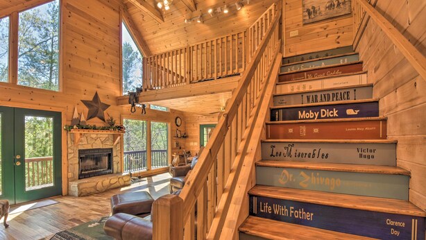 A wooden cabin interior with a fireplace, a staircase with books, and a view of the outdoors. - Dreamy Ellijay Resort Cabin w/ Game Room & Decks! (Ellijay)