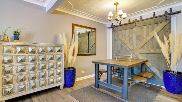 Une pièce avec un plancher en bois, un tapis bleu tissé, une table en bois avec deux chaises, un pot bleu tissé contenant de l’herbe des pampas et un meuble blanc orné de miroirs décoratifs.