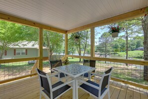 A covered outdoor patio with a table and chairs, surrounded by a wooden deck and railings, overlooking a green landscape and a body of water. - Waterfront Lake Sinclair Home: Dock + Grill! (Eatonton)