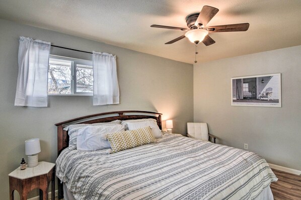 A bedroom with a bed, a ceiling fan, a window with curtains, a bedside table with a lamp, and a framed picture on the wall.