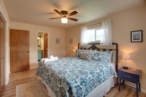 Une chambre à coucher comprenant un lit, une table de chevet, une lampe, un ventilateur de plafond, une fenêtre avec des rideaux et une porte donnant sur une salle de bain.