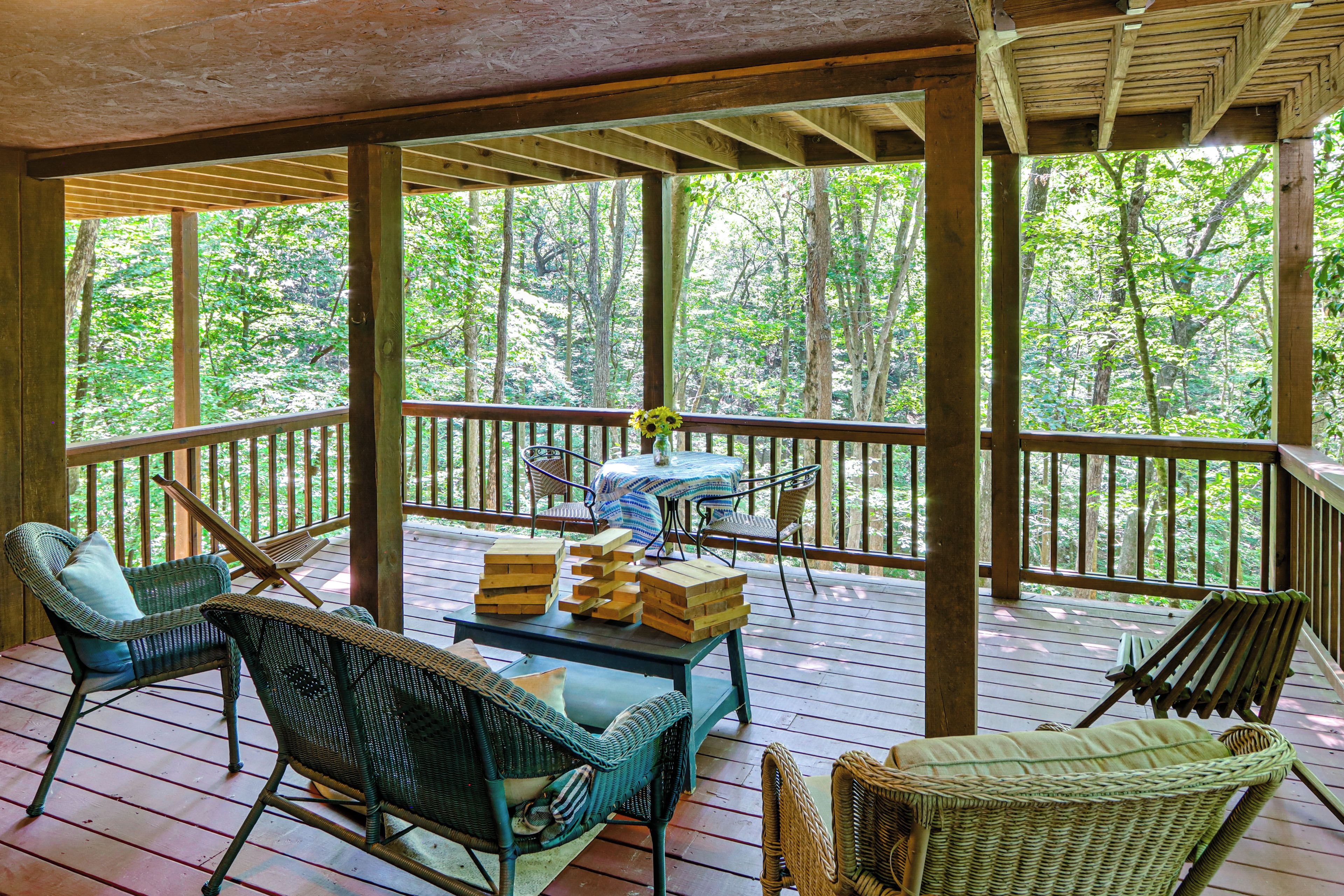 A covered outdoor deck with wicker furniture, a table with a vase of flowers, and a view of a forest.