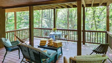 A covered outdoor deck with wicker furniture, a table with a vase of flowers, and a view of a forest.