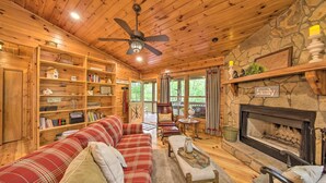 Interior acogedor de una cabaña con techo de madera, chimenea, sofá rojo a cuadros y estantería llena de libros y objetos decorativos.