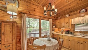 Interior de una cabaña de madera con una mesa redonda para comer, sillas, una araña y una zona de cocina con armarios de madera y una ventana con vista a una terraza.