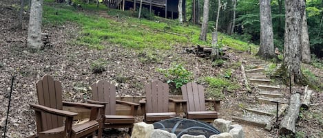 Ein bewaldetes Gebiet mit einer Holzterrasse, Adirondack-Stühlen und einer Feuerstelle aus Stein.