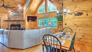 A cabin interior with a stone fireplace, a large window, a ceiling fan, and a dining table with chairs. - Serene Blue Ridge Cabin ~ 3 Mi to Downtown! (Blue Ridge)