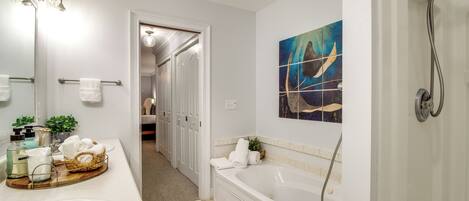 A bathroom with a large bathtub, a sink, and a mirror. There is a towel rack with towels and a basket with toiletries. A painting is on the wall above the bathtub.