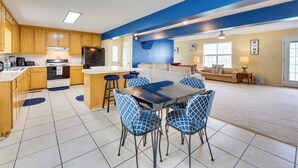 A spacious living area with a large dining table and blue patterned chairs, a kitchen with wooden cabinets, and a blue accent wall.