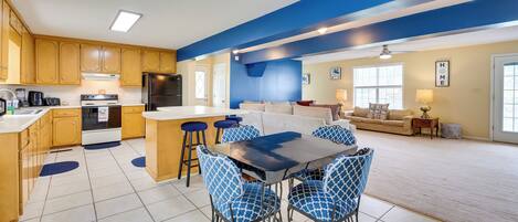 Amplia sala de estar con una gran mesa de comedor y sillas azules estampadas, una cocina con gabinetes de madera y una pared azul de acento.