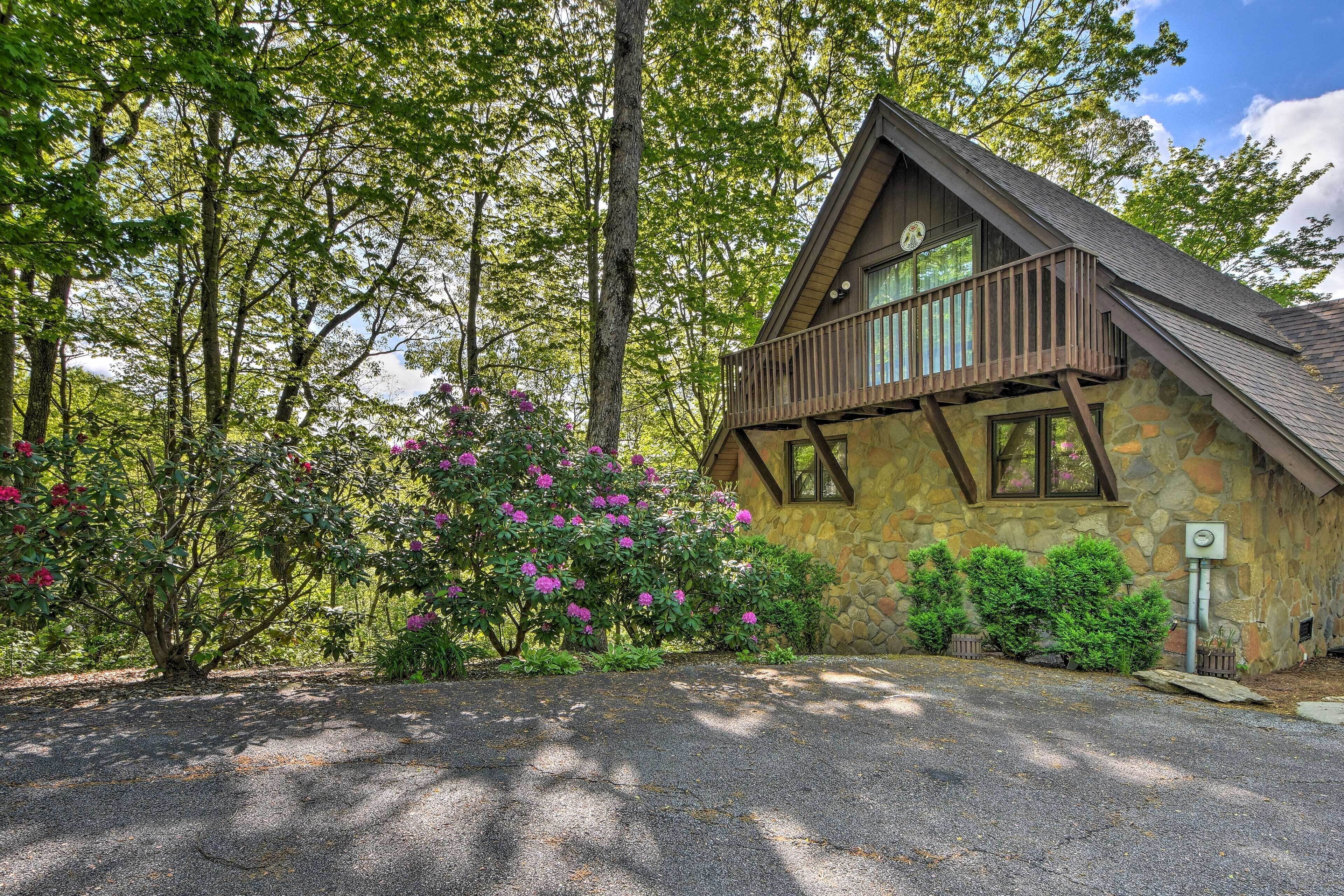 A stone house with a wooden balcony, surrounded by trees and flowering bushes.