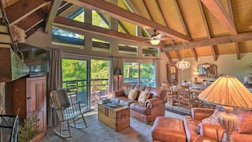 A spacious living area with a high ceiling, wooden beams, a sofa set, a rocking chair, and a dining area with a chandelier.