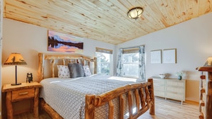 Una cama con estructura de madera y colcha estampada, una mesita de noche con lámpara, un aparador y un cuadro de un paisaje montañoso.