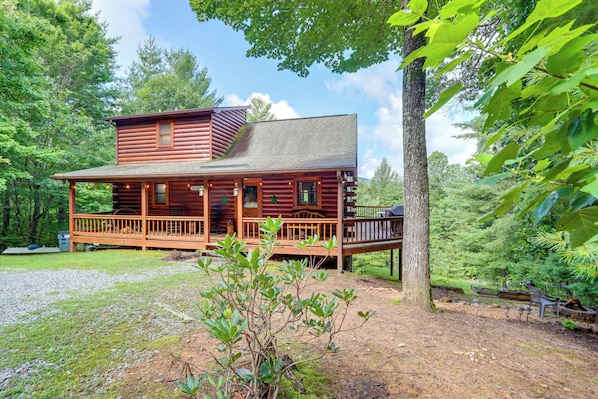 A log cabin with a porch, surrounded by trees and a gravel driveway.