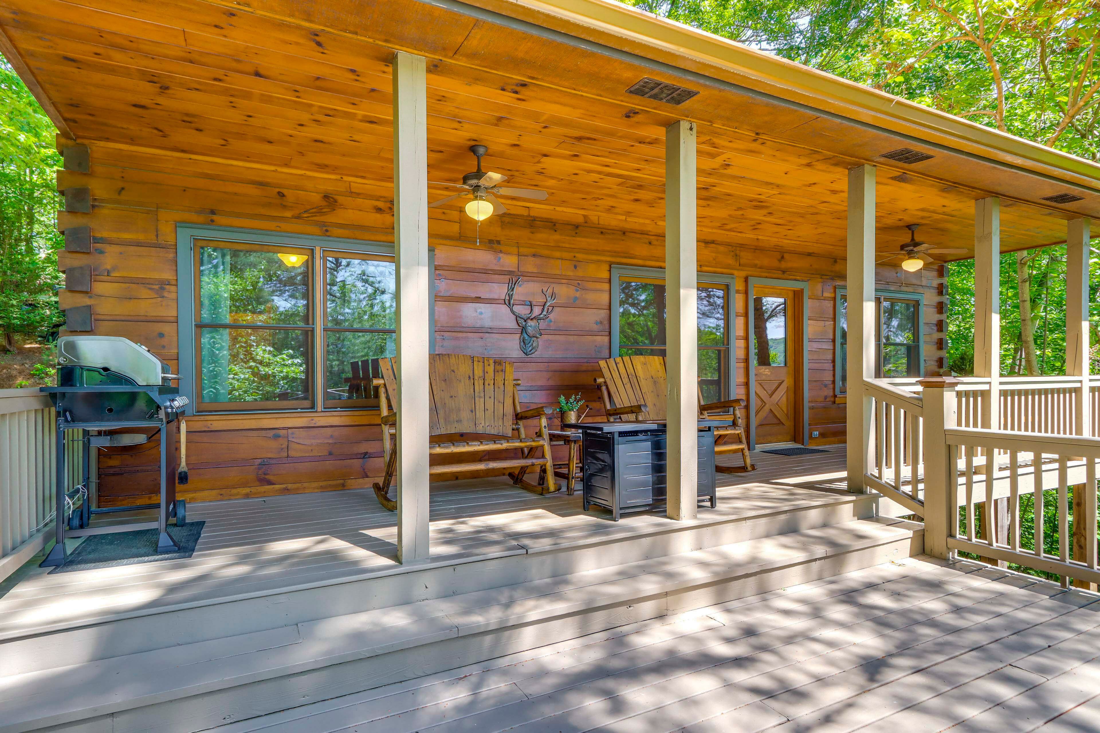 A wooden cabin with a porch, a ceiling fan, a grill, and a deer head wall decoration.