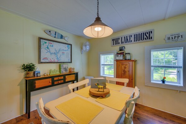 Un coin repas avec une table, des chaises, une carte accrochée au mur, une enseigne indiquant « THE LAKE HOUSE », et une fenêtre donnant sur un paysage verdoyant.