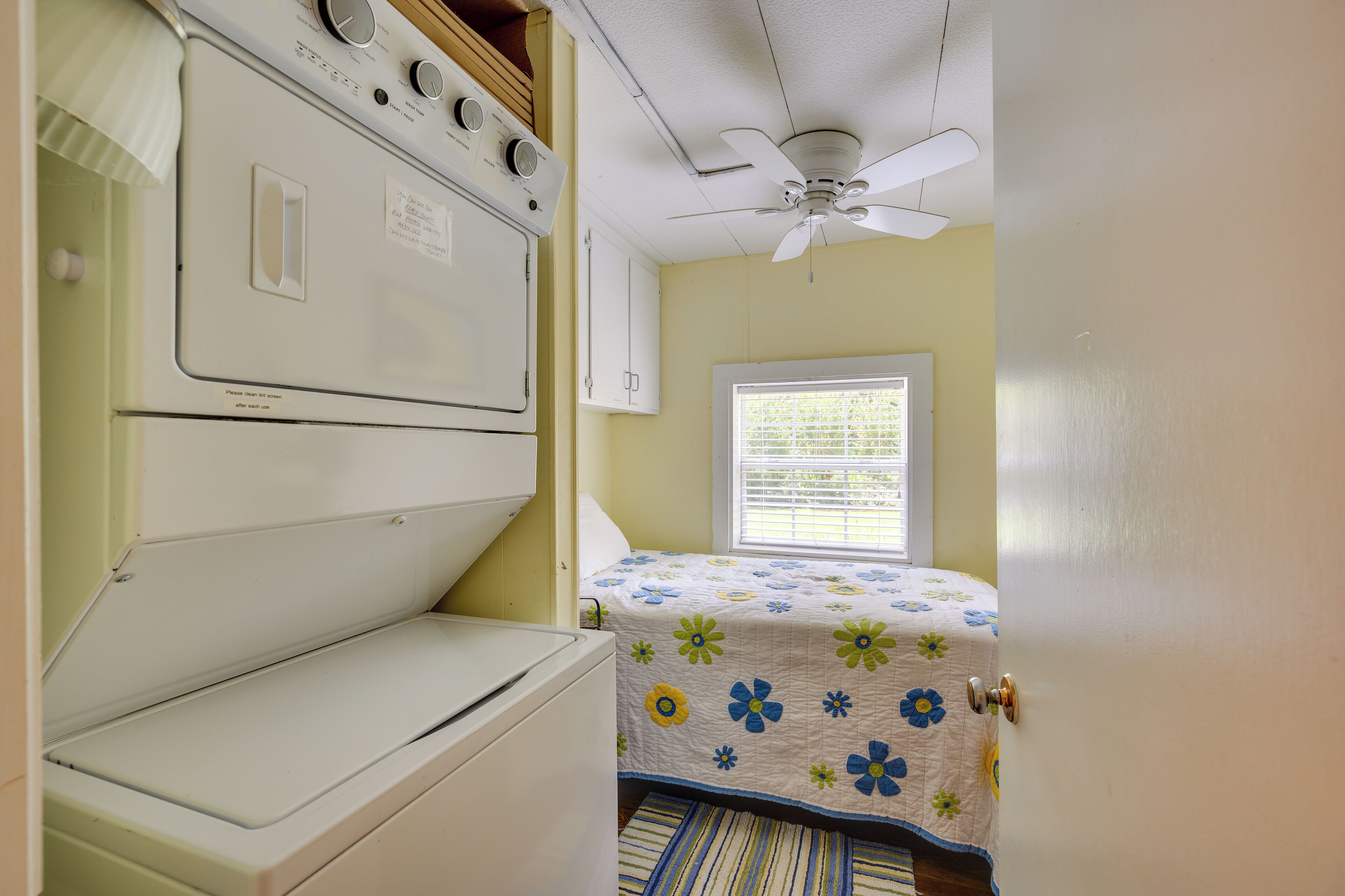 A compact room with a bed, a ceiling fan, a window with blinds, and a washing machine.