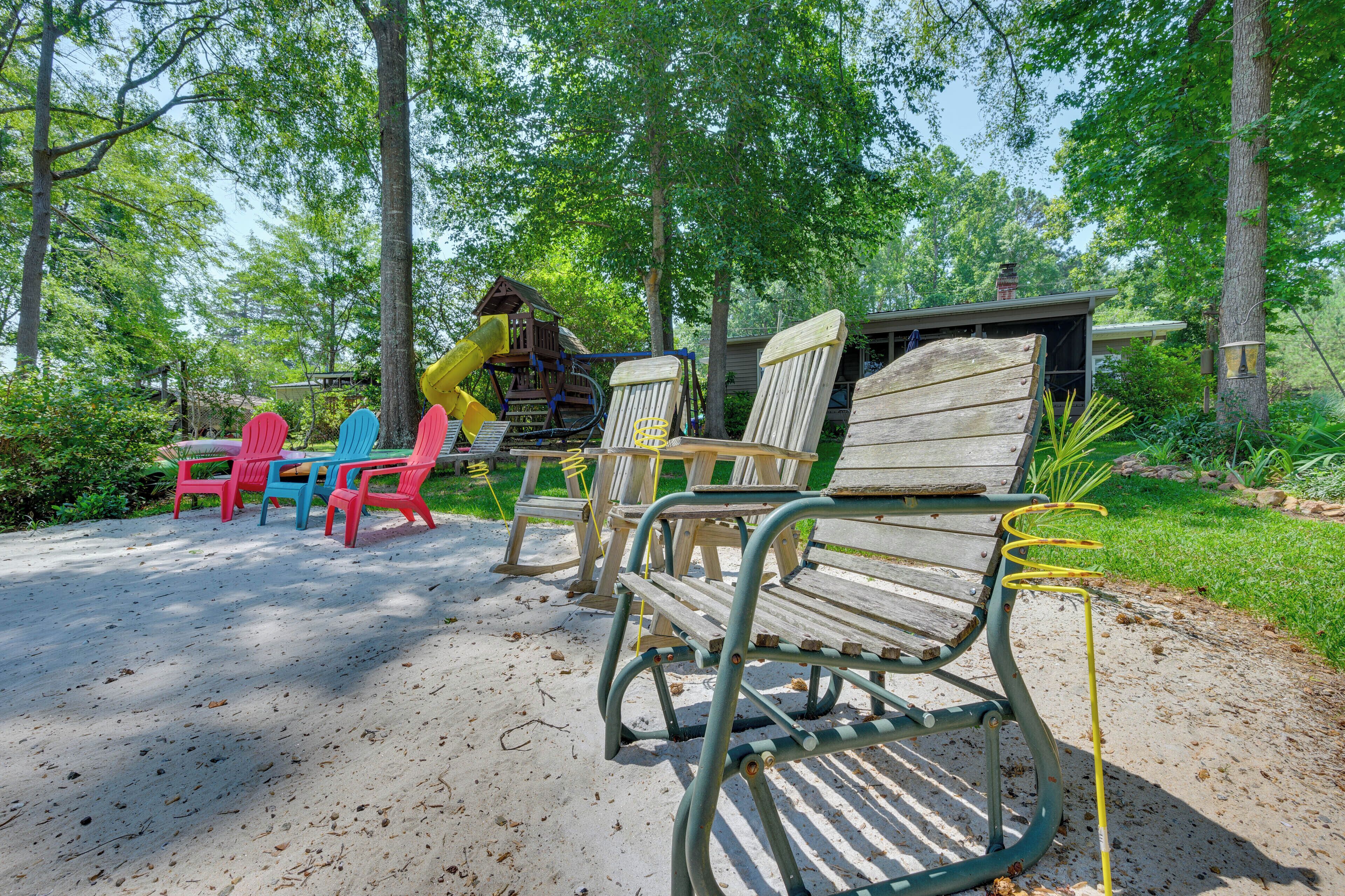 A wooden bench and colorful Adirondack chairs in a sandy outdoor area with trees and a wooden structure in the background.