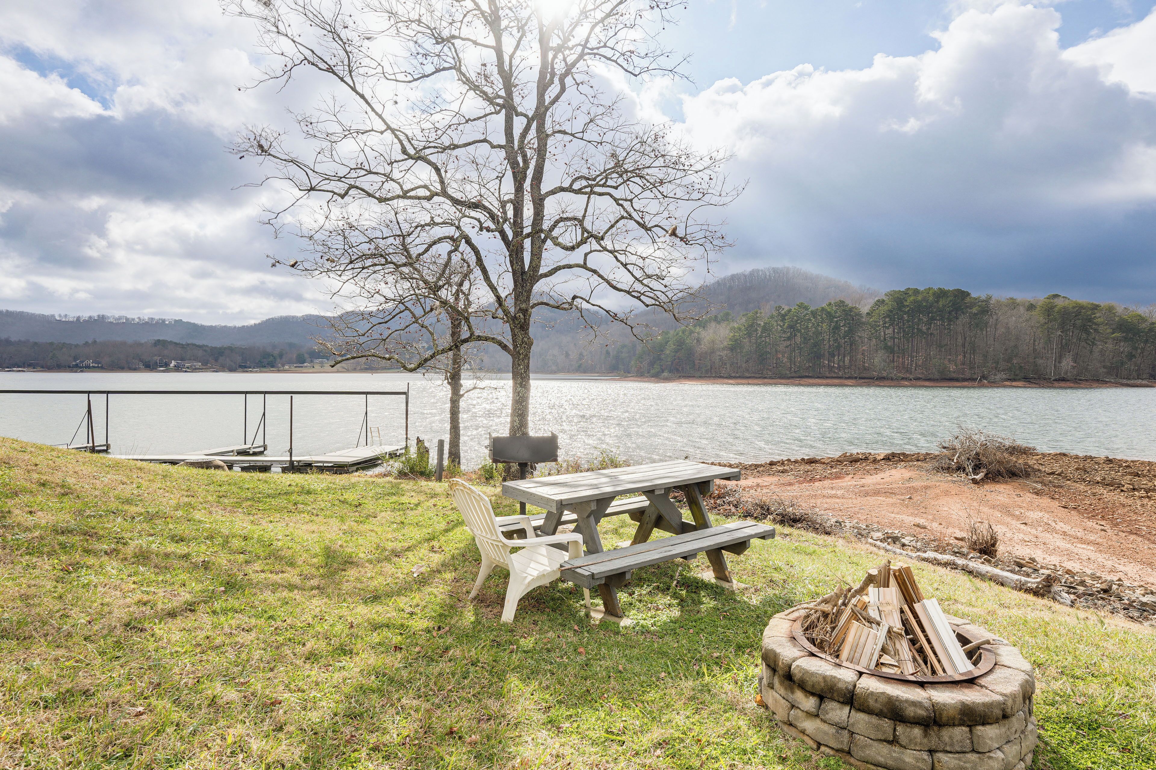 A lakeside picnic area with a wooden table and benches, a fire pit, and a dock with a boat.