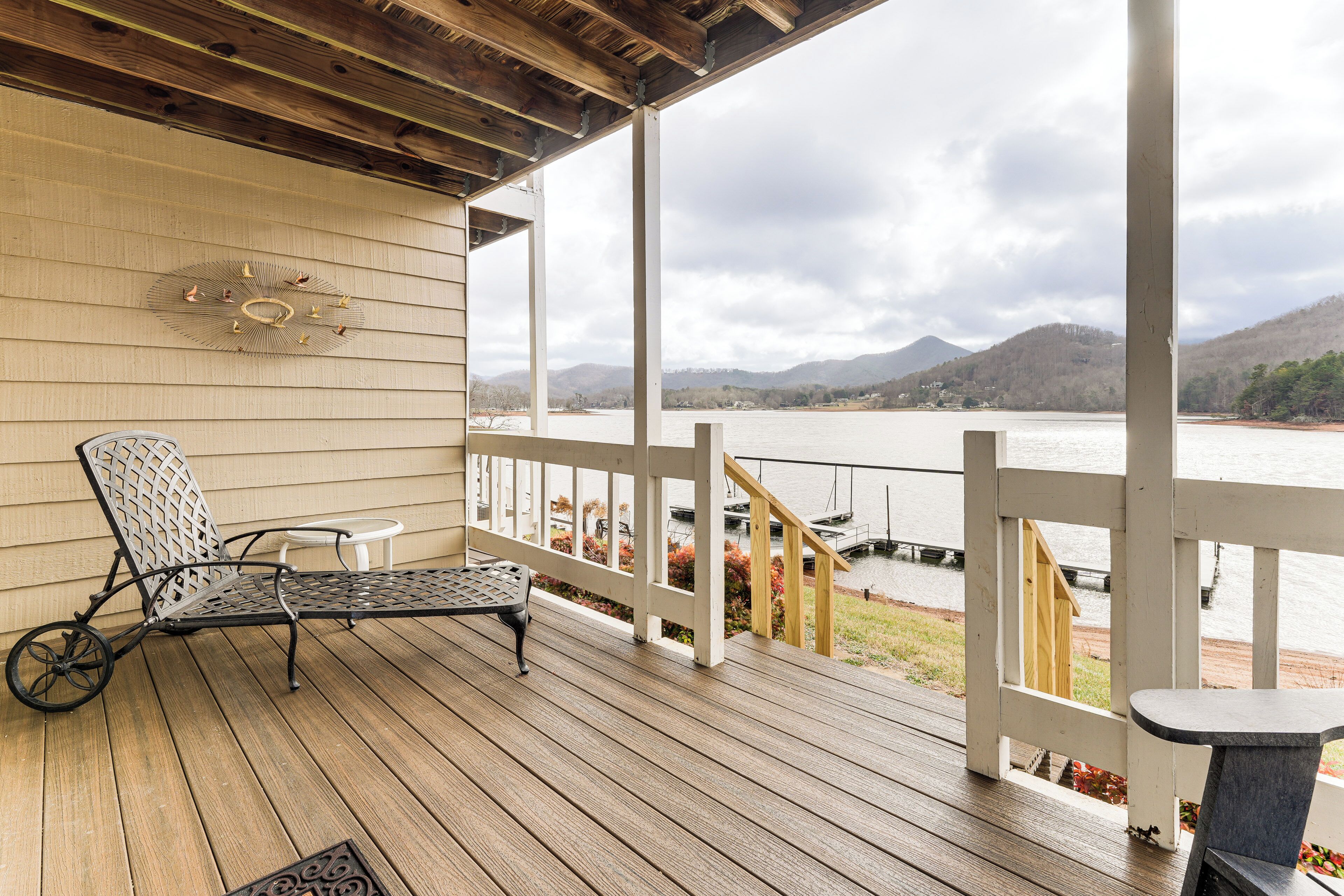 Eine Holzterrasse mit einem Metallstuhl und einem kleinen Tisch, mit Blick auf einen See mit Booten und Bergen im Hintergrund.
