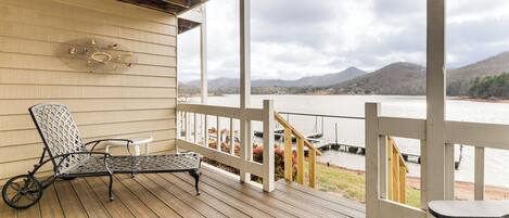 A wooden deck with a metal chair and a small table, overlooking a lake with boats and mountains in the background.