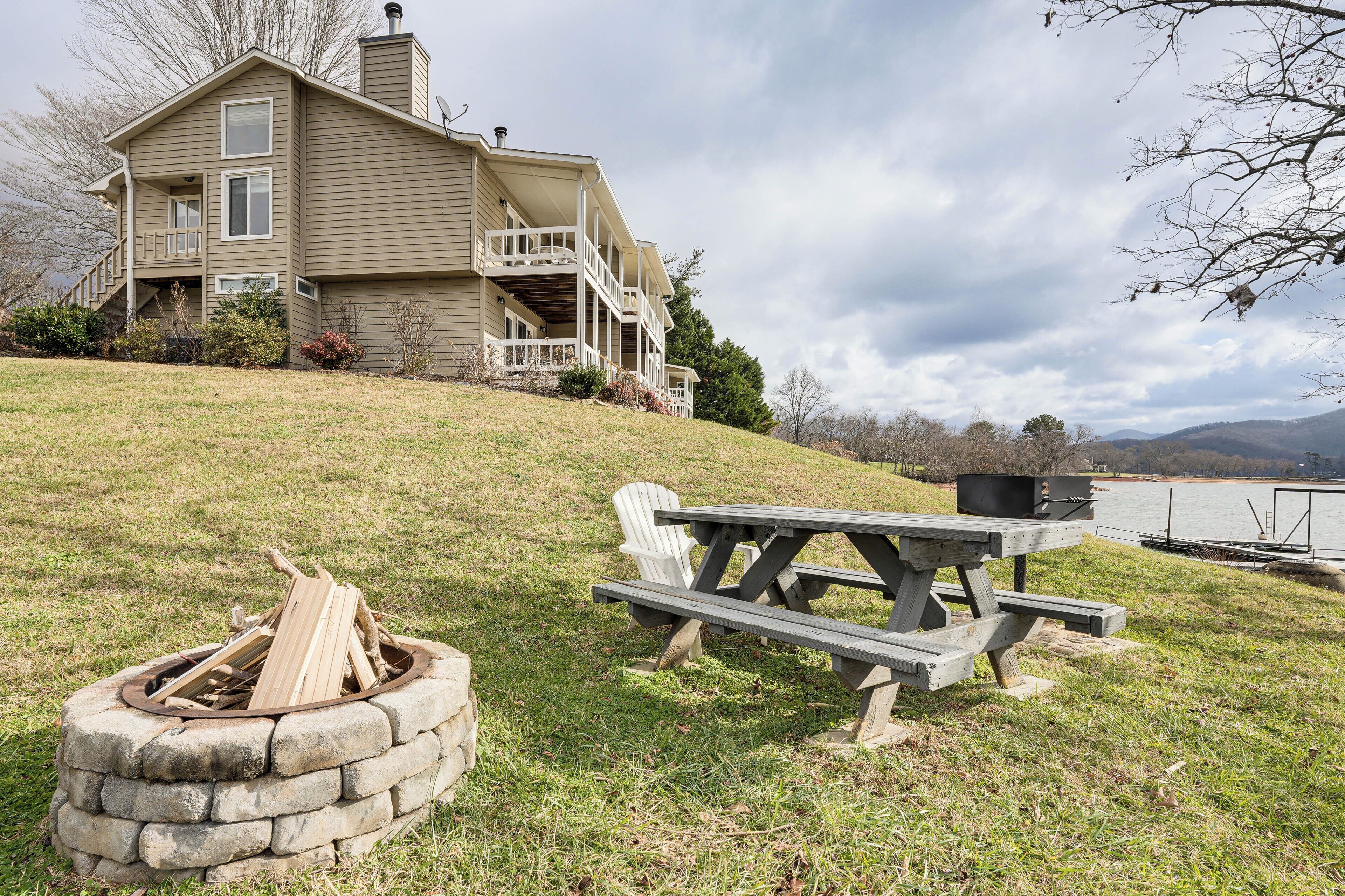 A house with a porch, a stone fire pit, and a picnic table by a lake.