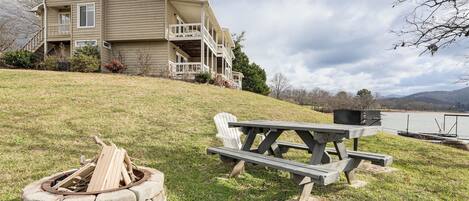 A house with a porch, a stone fire pit, and a picnic table by a lake.