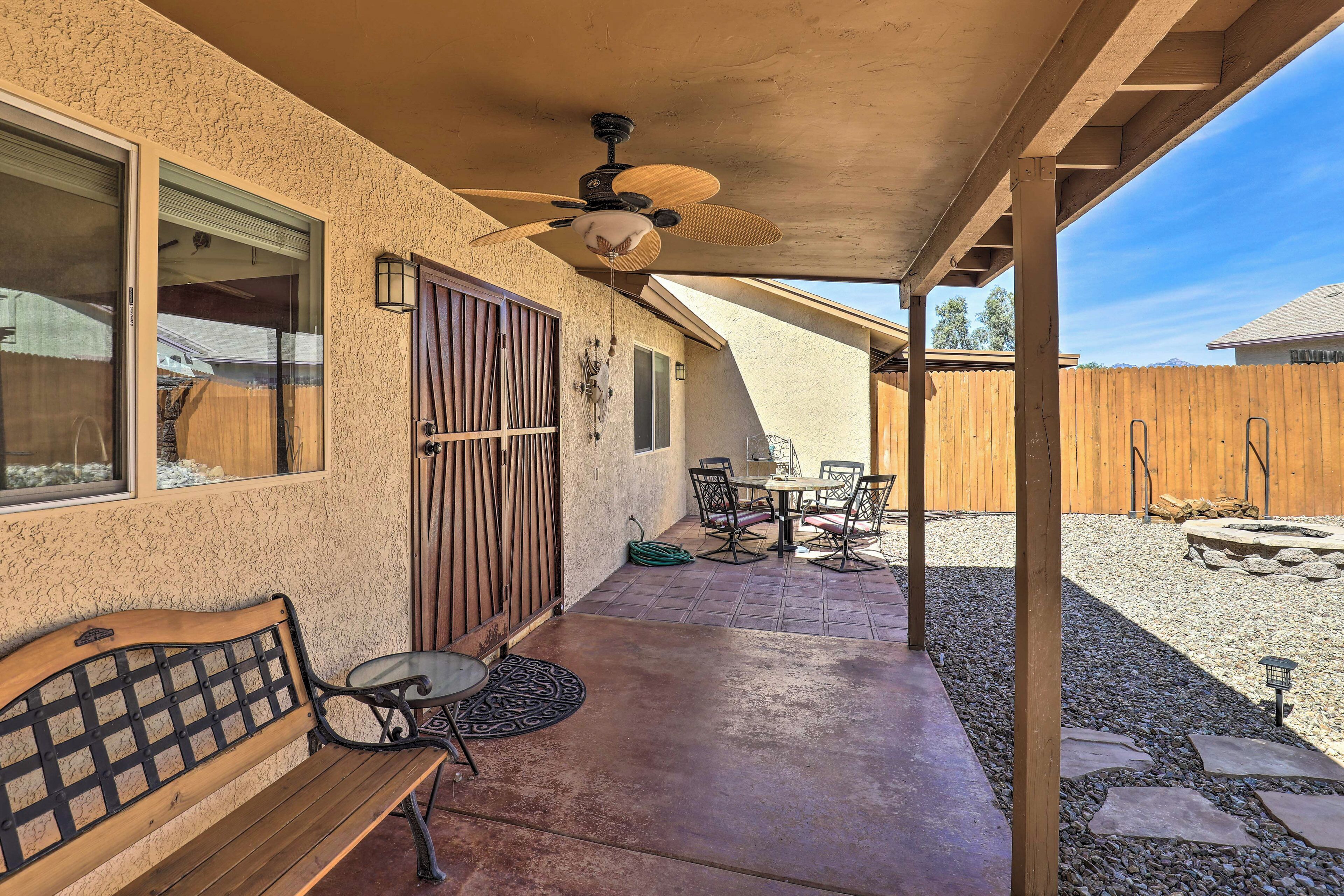 A covered patio with a ceiling fan, a wooden bench, a table with chairs, and a view of a backyard with a wooden fence and a stone fire pit.