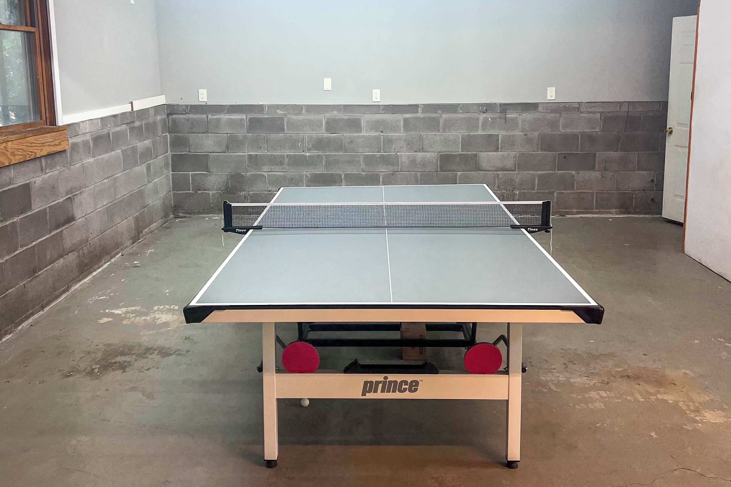 Een tafeltennistafel met een net van het merk Tinos, geplaatst in een ruimte met een betonnen vloer en een bakstenen muur.
