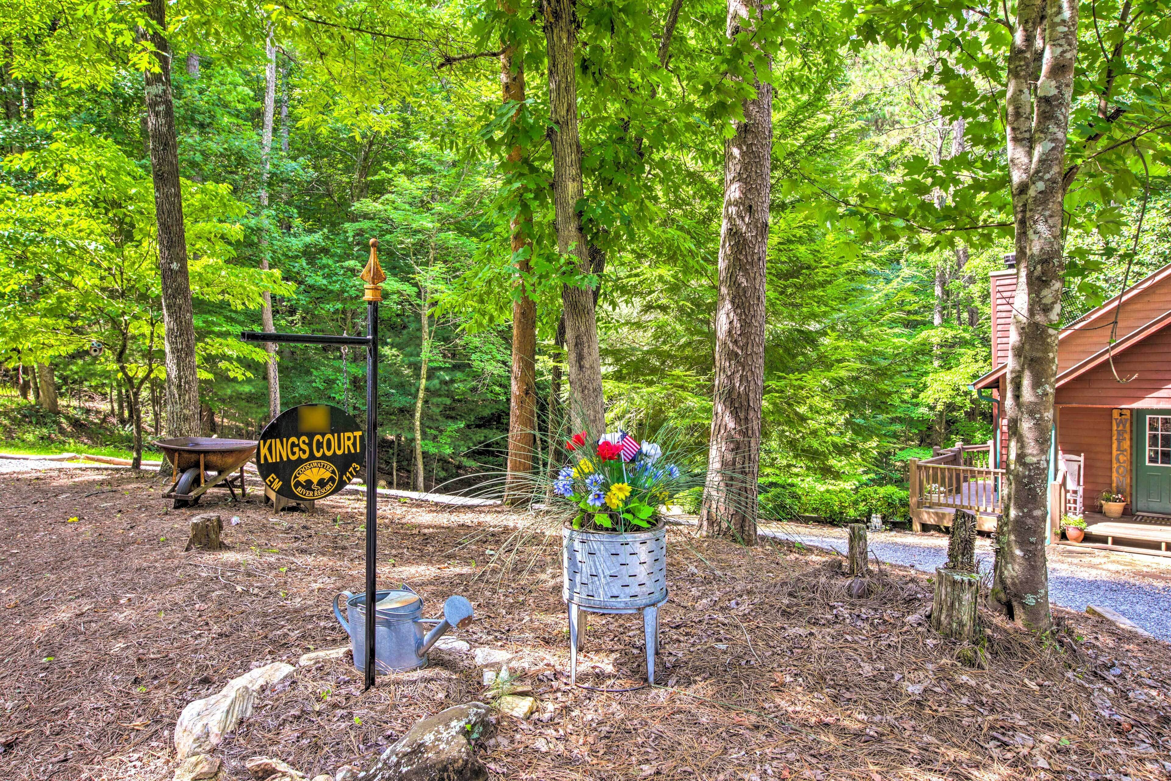 A rustic cabin with a wooden deck, surrounded by a forest. A metal sign with "Kings Court" is visible.