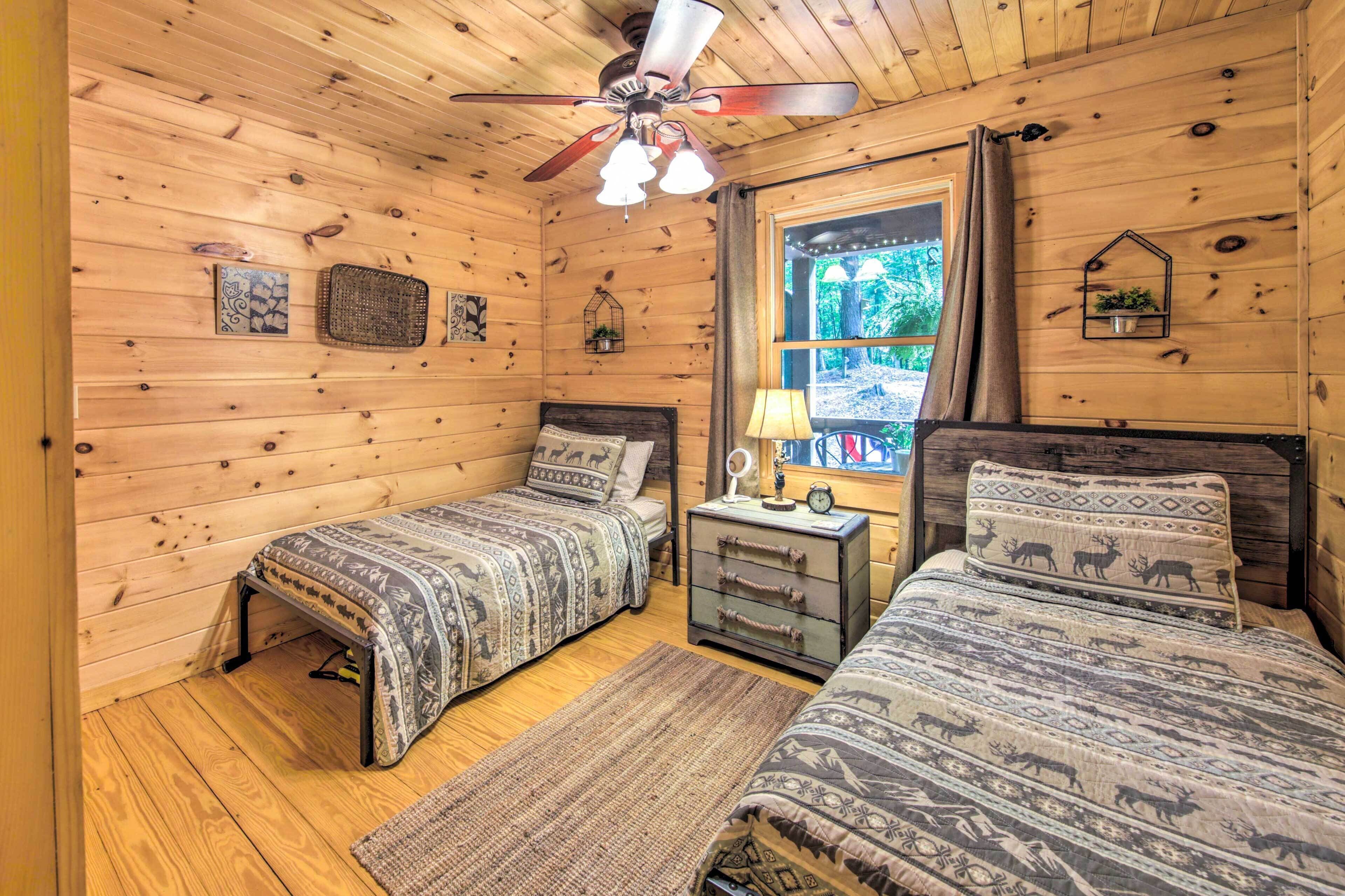 A cozy cabin room with two beds, wooden walls, a ceiling fan, and a window with curtains.