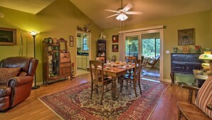 A dining area with a wooden table, chairs, and a patterned rug. There is a ceiling fan, a fireplace, and a view of the outdoors through a glass door.